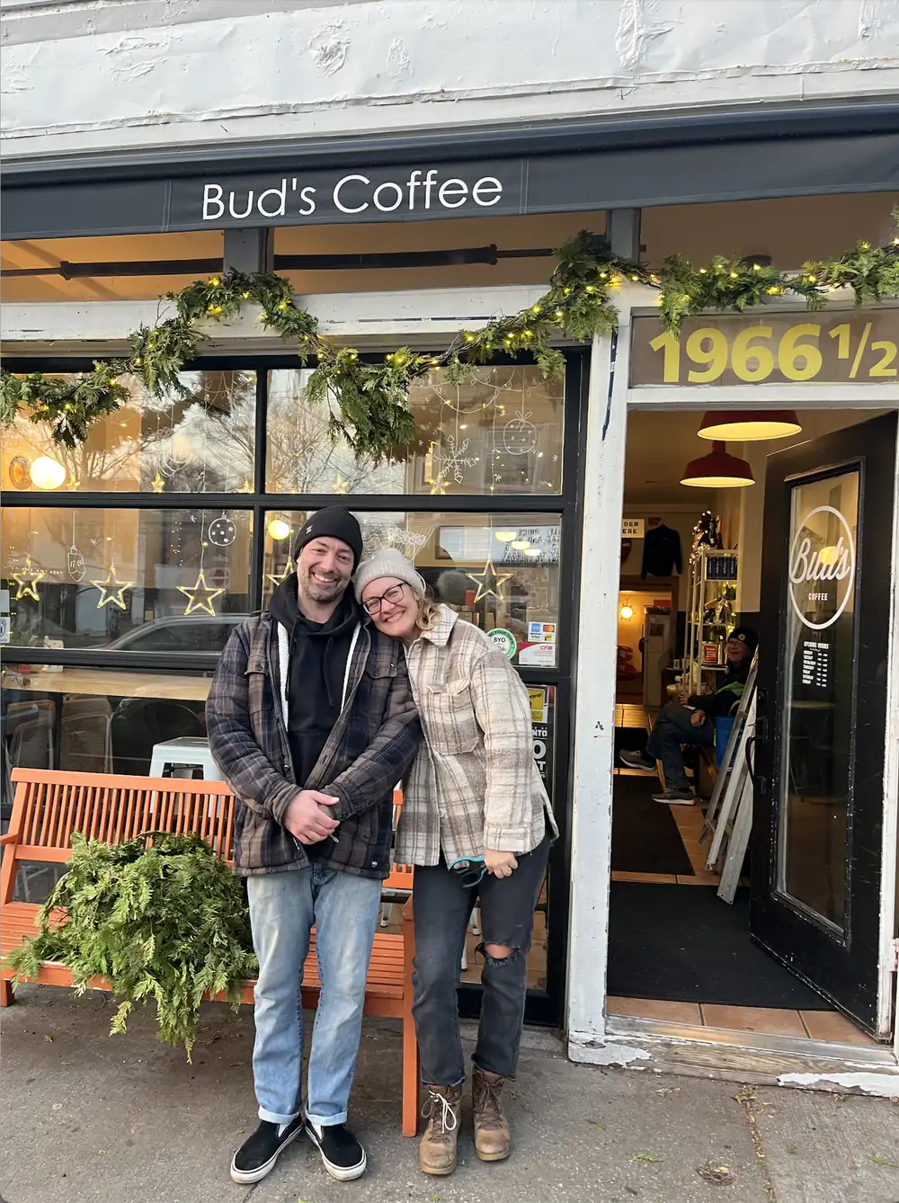 Sarah and Ken, the owners, smiling arm in arm outside the open door of Bud's Coffee, with garland and lights over the window