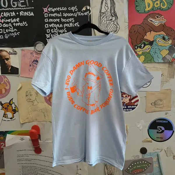 A grey T-shirt with an orange Bud's "damn good coffee" print, hung on the sticker-covered wall