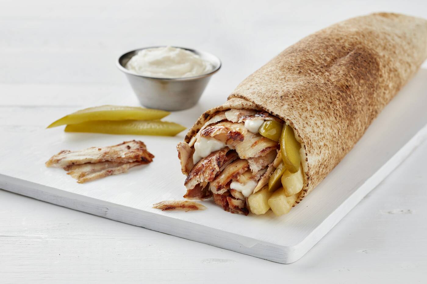 A chicken shawarma wrap with pickles next to a cup of garlic sauce