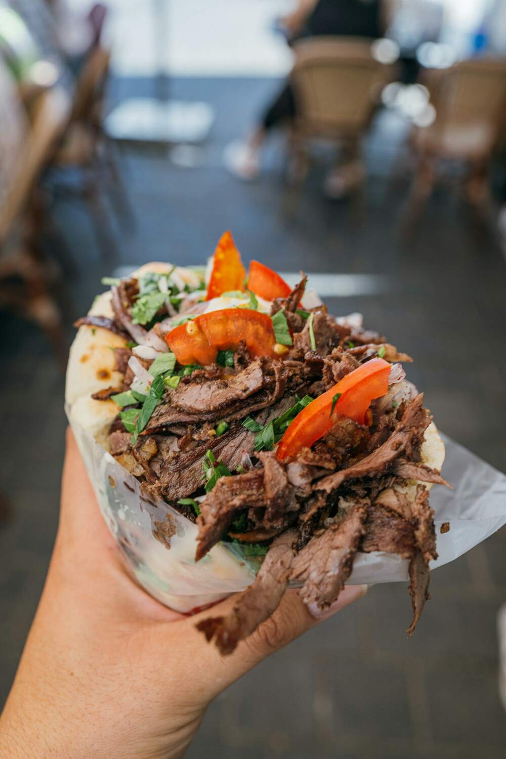 A hand holds a pita piled with sliced beef, tomato and parsley