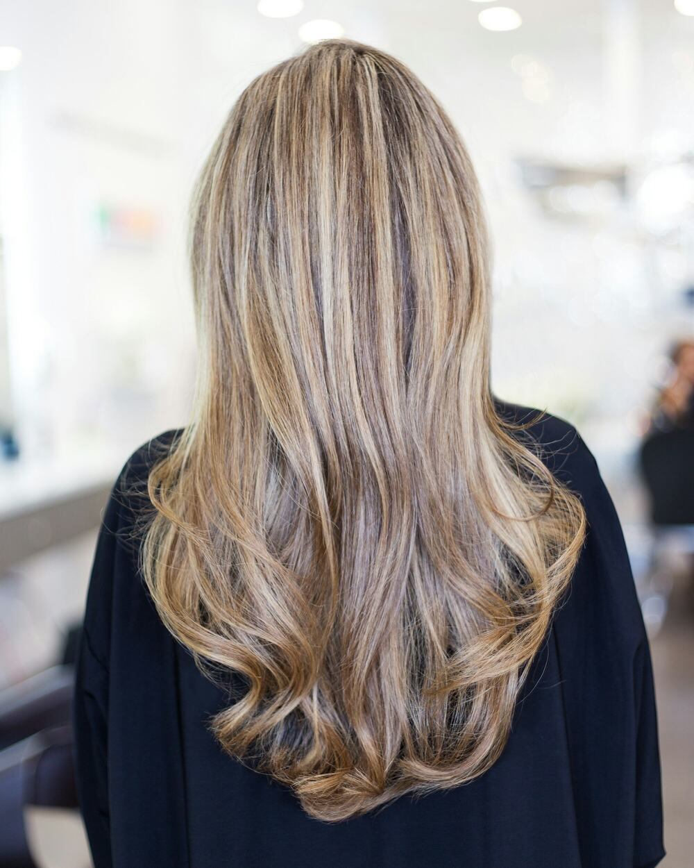 Long, softly curled hair with blonde highlights, seen from behind in a salon