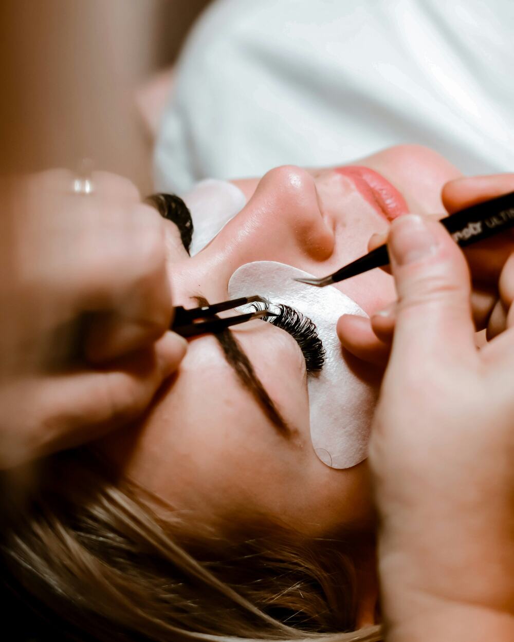 An esthetician applying eyelash extensions with tweezers to a client lying back with eyes closed