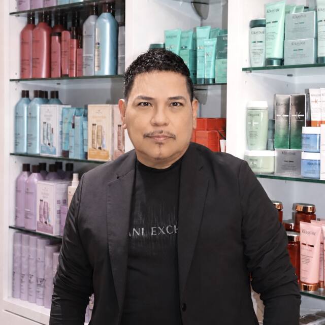 Rigo, Sr. Stylist, in front of the salon’s product wall
