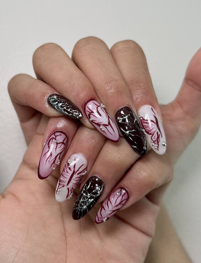 Almond nails alternating deep burgundy with silver chrome and milky white with hand-drawn burgundy hearts and wings, set with silver beads