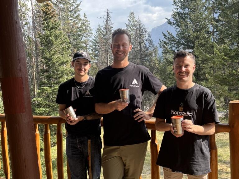 Three people on a wooden porch among pine trees, each holding a Muuse cup