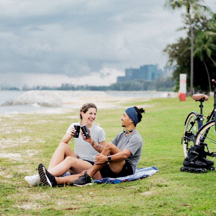 Two friends on a picnic blanket by the water, toasting with Muuse cups