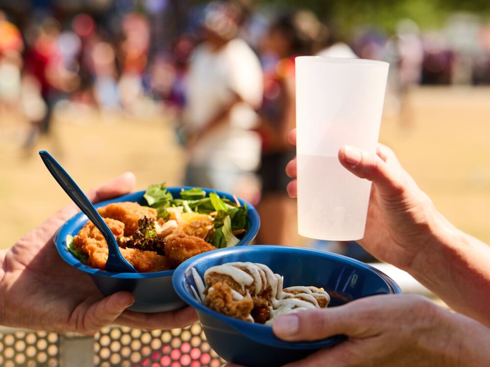 Hands holding blue reusable bowls of food and a reusable cup at an outdoor event
