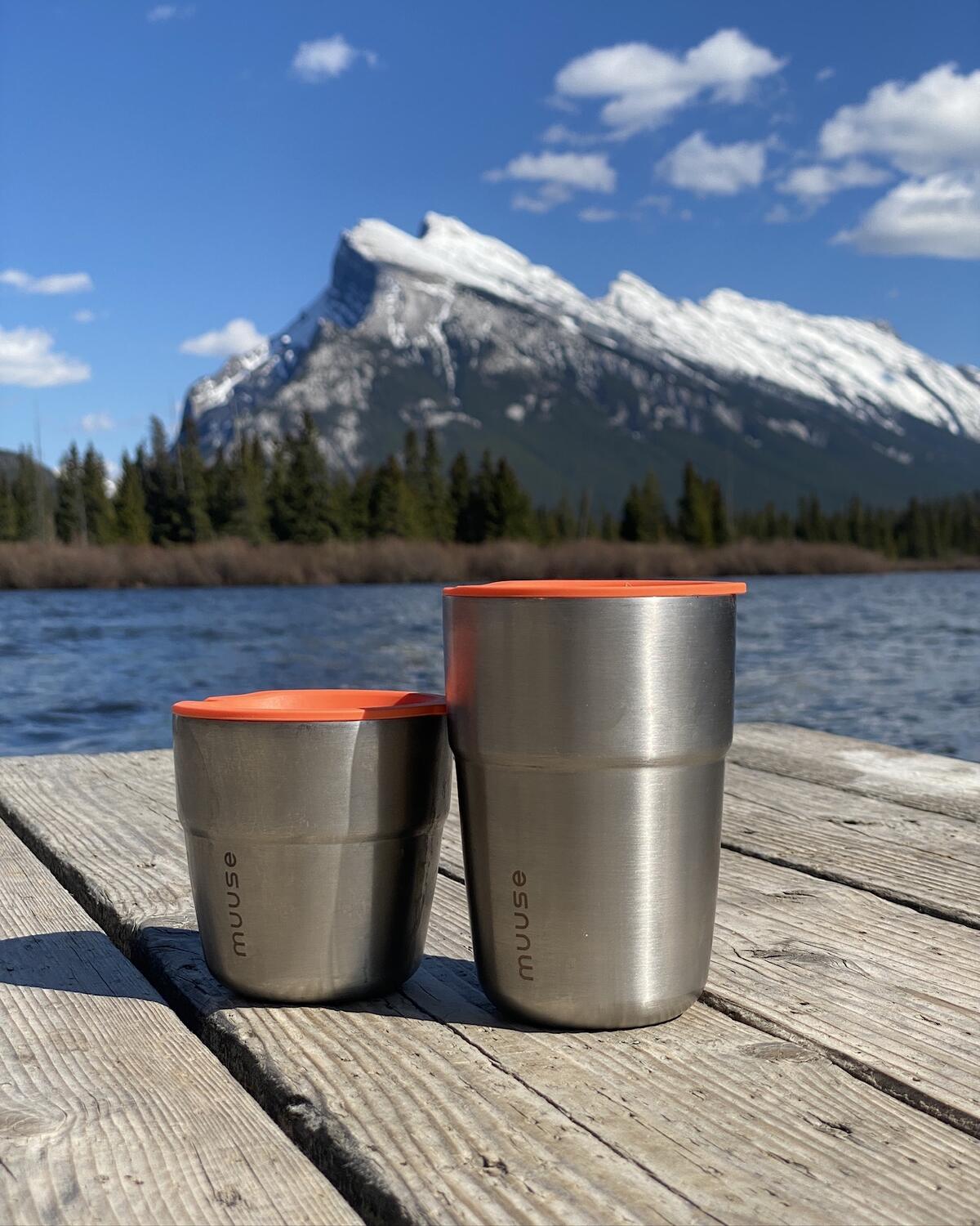 Two stainless steel Muuse cups with orange lids on a wooden dock, a snowy mountain behind