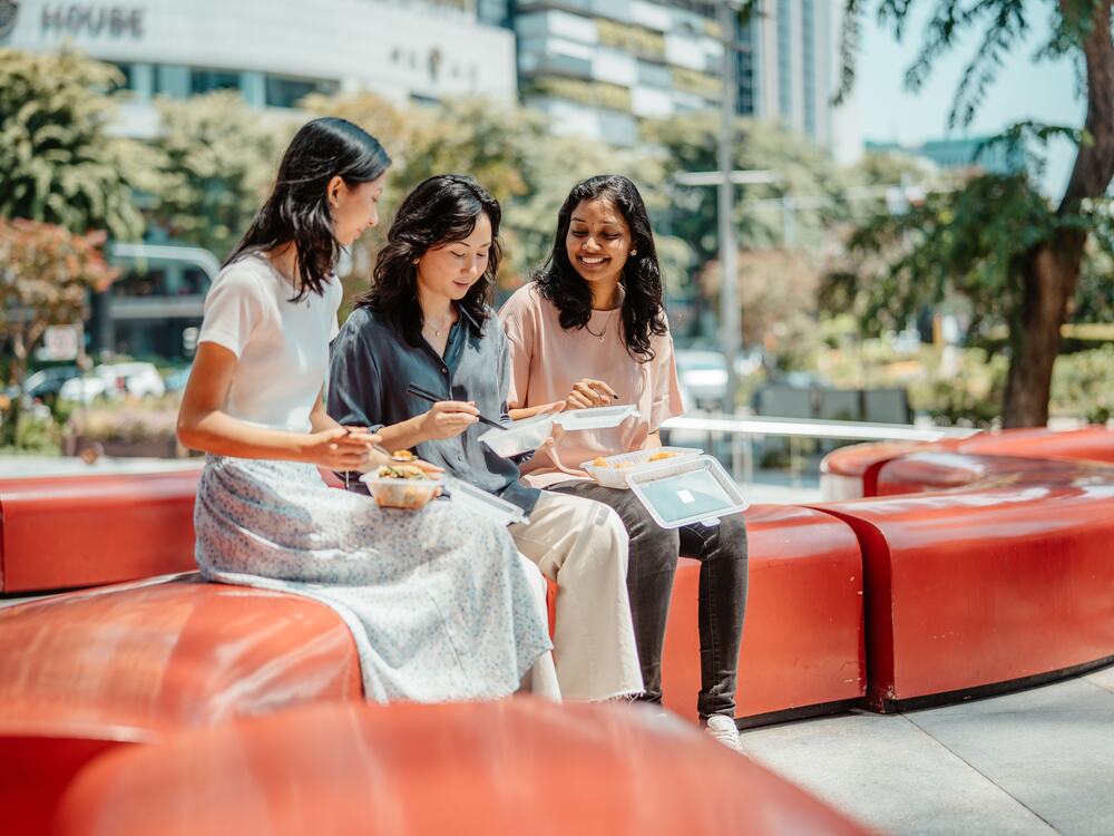 Three friends on a red outdoor sofa sharing lunch from reusable Muuse containers