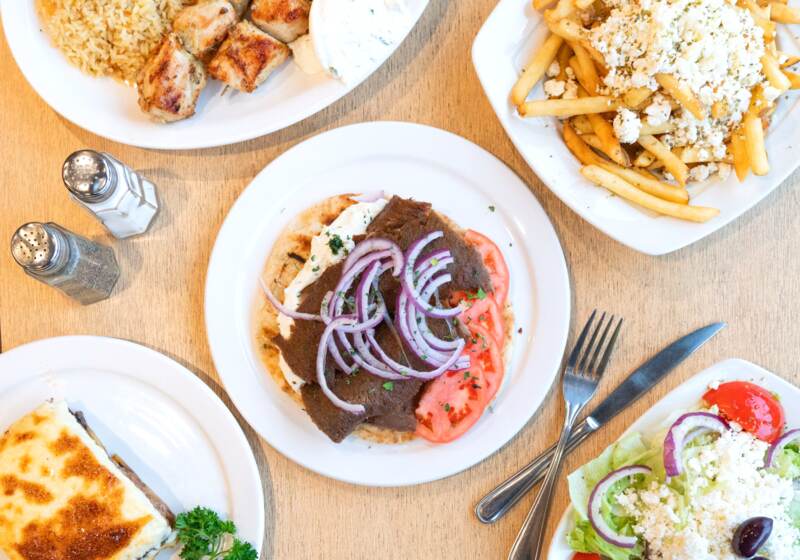Overhead view of a table at Souvlaki Hut: gyros on pita with red onion and tomato, chicken souvlaki with rice, Greek fries and a Greek salad