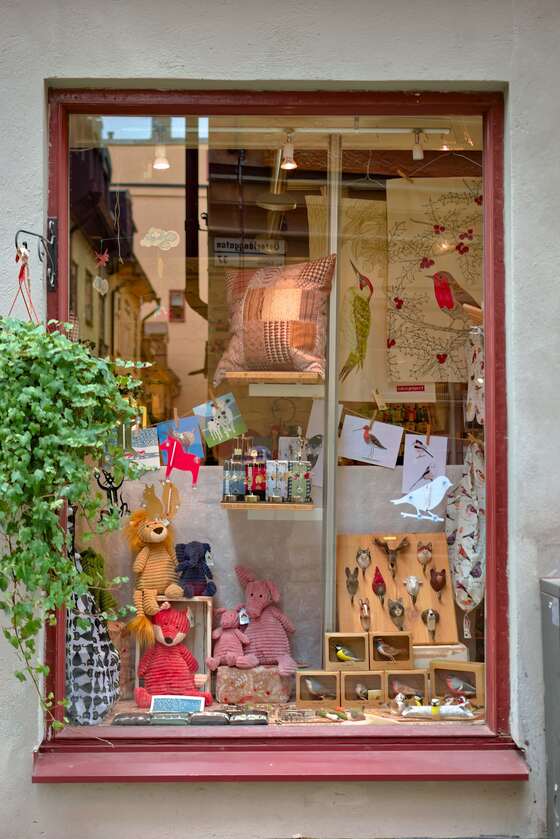 A small shop window with a red frame, filled with soft toys, cards, cushions and little wooden boxes, with a trailing plant beside it