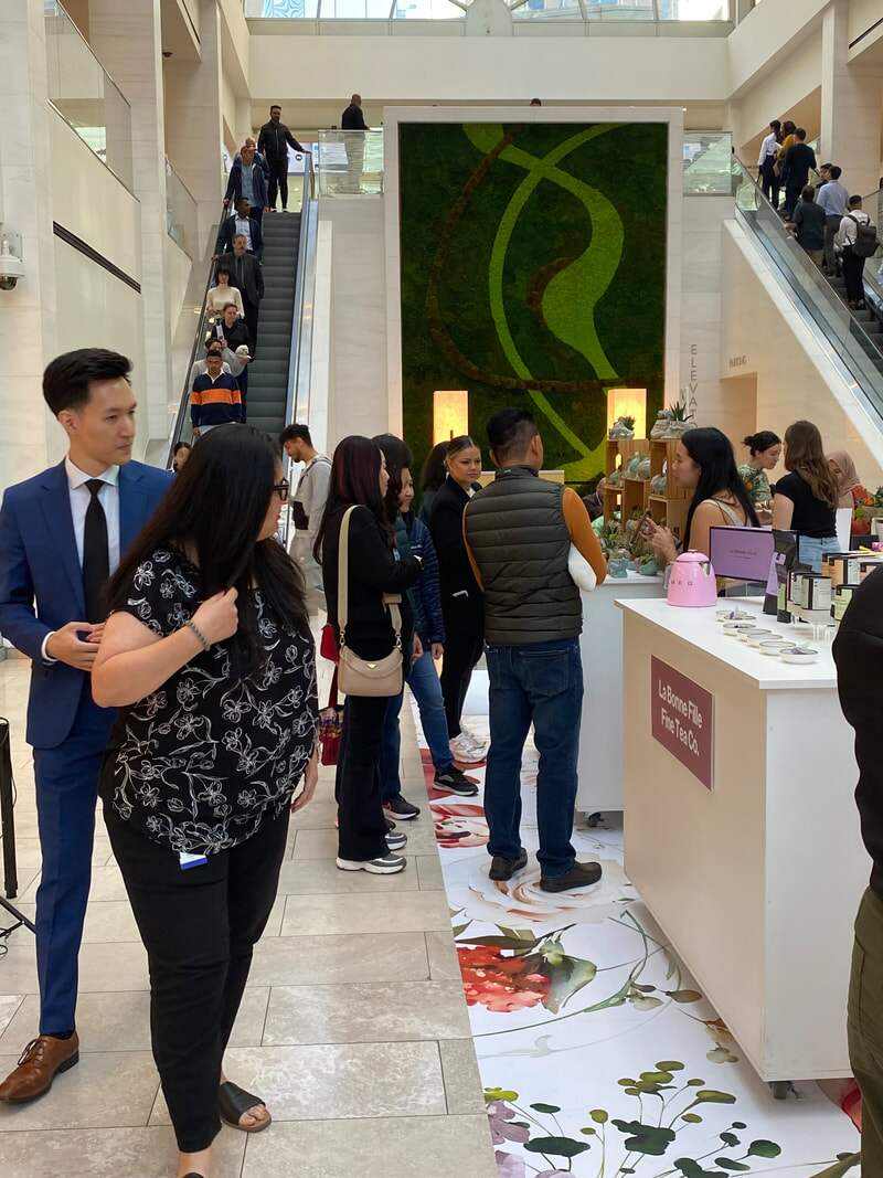Shoppers gather at vendor tables in a bright office-tower atrium beside a living green wall