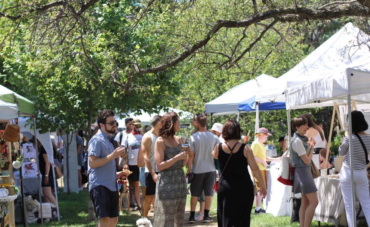 Shoppers stroll between white vendor tents under the trees at an outdoor Toronto Artisan Market