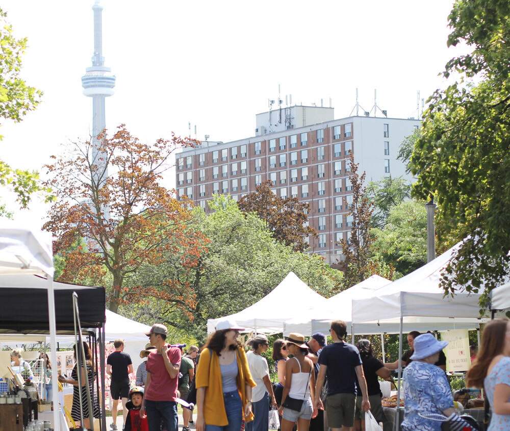 Shoppers walk past market tents in a Toronto park, with the CN Tower rising behind the trees